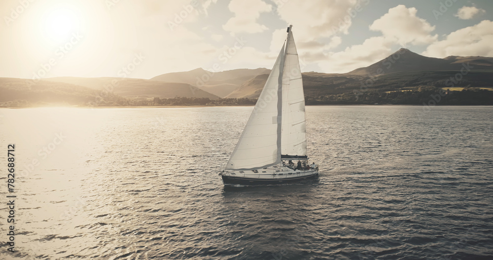 Sun seascape with white sailboat aerial. Mesmerezing landscape with sail boat at summer. Sunlight over sail boat at ocean bay. Sea shore of Arran Island with vessel. Serene water scenery drone shot