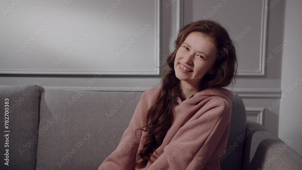 A happy european or american young lady sits on the sofa, squeezes shoulders, smiles and laughs. A girl teenager model in a joyful mood and positive emotions poses and looks at the camera in a playful A happy european or american young lady sits on the sofa, squeezes shoulders, smiles and laughs. A girl teenager model in a joyful mood and positive emotions poses and looks at the camera in a playful