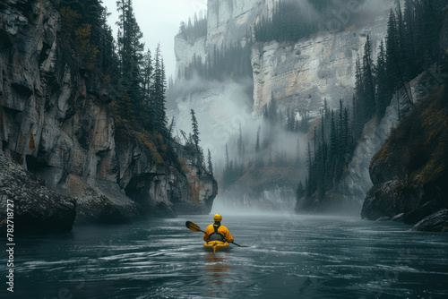 A man in a yellow jacket kayaks down the middle of steep mountains, with dark green trees and misty river water. photo