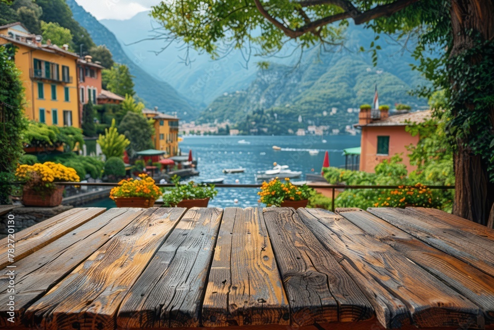 An empty wooden tabletop has a blurred background of a village by the lake, featuring quaint buildings and cafe tables.