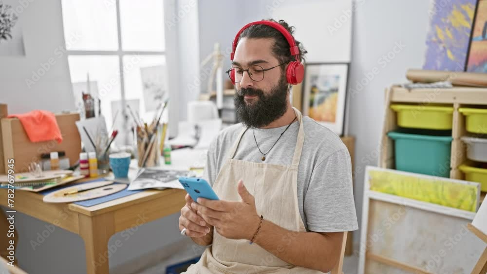 A bearded young man wearing headphones uses a smartphone in a creative art studio. A bearded young man wearing headphones uses a smartphone in a creative art studio.
