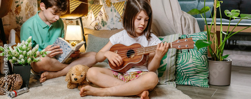 Banner of little girl playing ukulele while boy reading book on handmade teepee in living room. Children having fun in diy shelter tent in their house. Vacation camping at home or staycation concept. photo