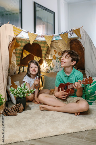 Happy children playing a ukulele and singing on handmade shelter tent in living room. Boy and girl laughing in a cozy diy teepee in house. Staycation and vacation camping at home concept.