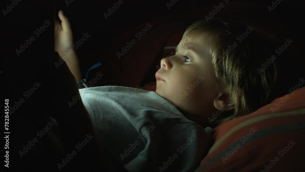 Young blonde boy engrossed in digital tablet under dim light at nighttime Young blonde boy engrossed in digital tablet under dim light at nighttime