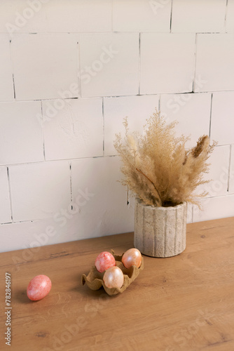 Three eggs are placed on a hardwood table next to a flowerpot filled with dried flowers and a twig. The wood flooring enhances the rustic feel of the setting © Маргарита Трушина