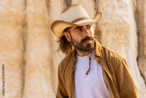 A man wearing a cowboy hat and a brown shirt is standing in front of a rock wall. He has a beard and a mustache