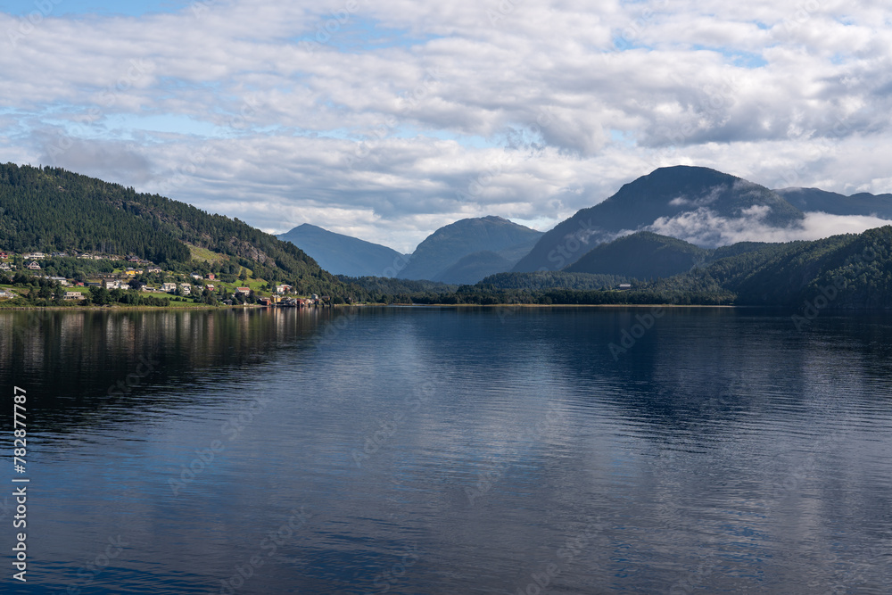 Calm waters of Stangvikfjorden mirror the sky, with the picturesque village of Kvanne nestled against a mountain, overlooking the Trollheimen mountains. Scandinavian travel destination in summer