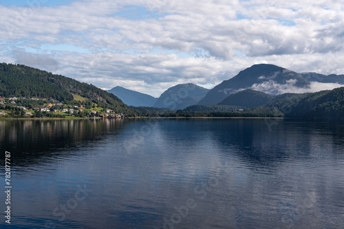 Calm waters of Stangvikfjorden mirror the sky, with the picturesque village of Kvanne nestled against a mountain, overlooking the Trollheimen mountains. Scandinavian travel destination in summer