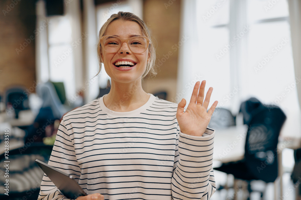 Female office worker holding digital tablet and waving Hi on camera standing on coworking background