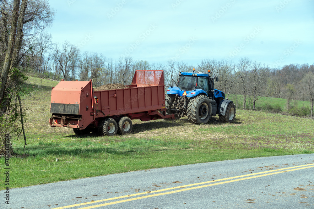 Fototapeta premium Blue tractor pulls a red fertilizer manure spreader over a green early spring agricultural field by a country road under a blue sky