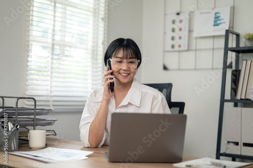 Cheerful businesswoman in white shirt using smartphone with laptop and documents on her office desk.