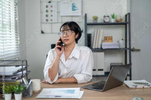 Cheerful businesswoman in white shirt using smartphone with laptop and documents on her office desk.