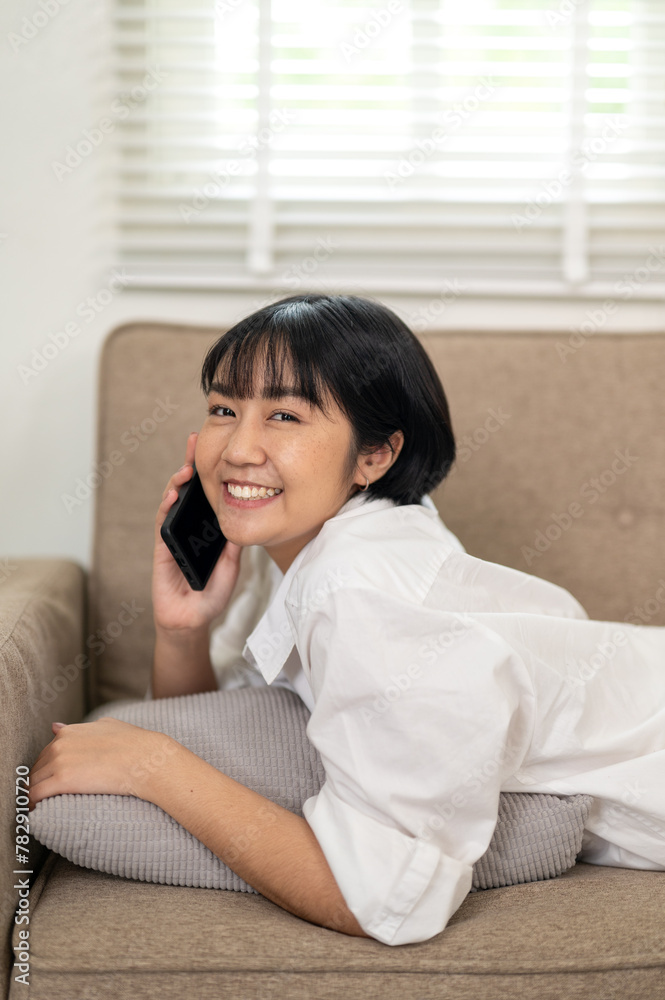 A smiling young woman enjoys a casual conversation on her smartphone while relaxing comfortably on a couch in a well-lit living space. A smiling young woman enjoys a casual conversation on her smartphone while relaxing comfortably on a couch in a well-lit living space.