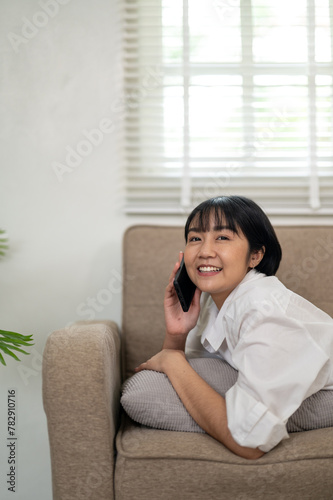 A smiling young woman enjoys a casual conversation on her smartphone while relaxing comfortably on a couch in a well-lit living space.