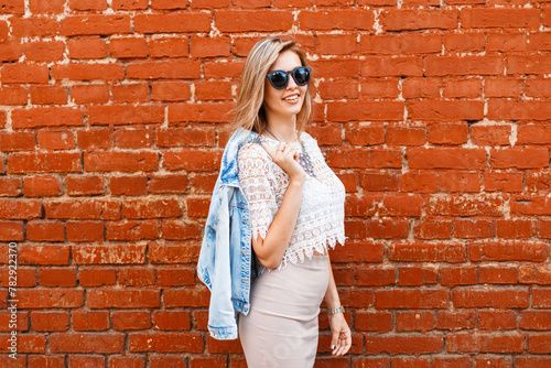 Happy fashion beautiful woman with cool sunglasses in fashionable lace top with denim jacket near red brick wall