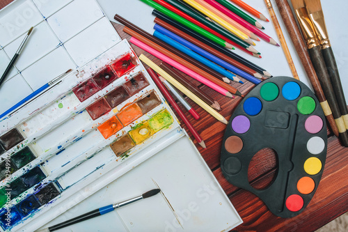 Top view of a multicolored watercolor palette, brushes, colored pencils and equipment for painting on white sheets of paper. Copy space, mockup.