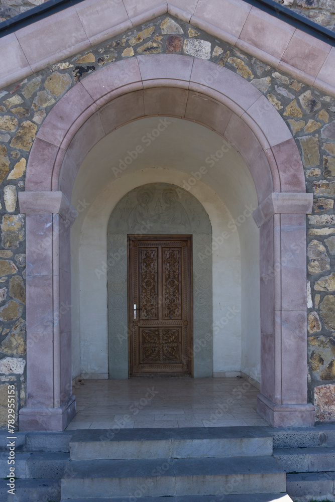 Carved wooden door of the Saint Peter and Paul monastery. Carved stone arch with patterns and bas-relief of saints. Brown stone arch and steps. View from outside of the church. Georgia