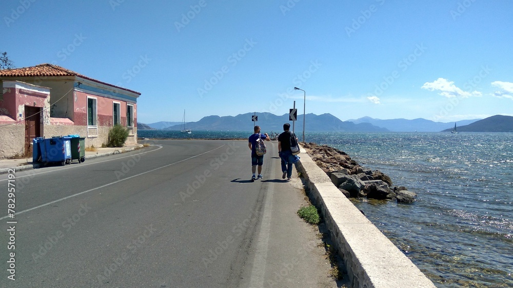 Two men walking on a road by the sea under a blue sky.