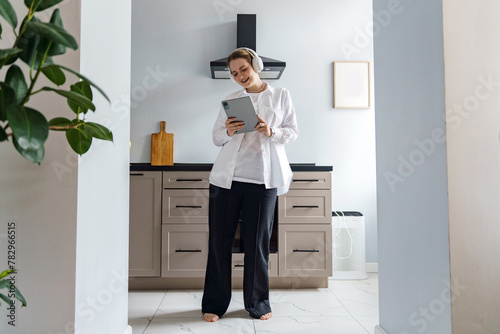 A relaxed professional in a modern kitchen enjoys a moment with her tablet and headphones, blending work and leisure.