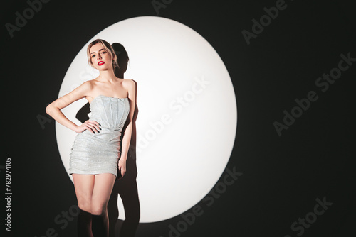 Young beautiful confident blond female in trendy evening silver dress. Sexy carefree woman posing near white wall in studio in a circle of light. Fashionable model with bright makeup and red lips