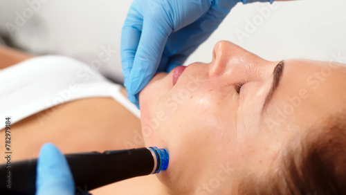 A middle-aged woman receives facial treatment in a professional beauty salon. A close-up cosmetologist manipulates a hydropeeling device to clean and rejuvenate facial skin.