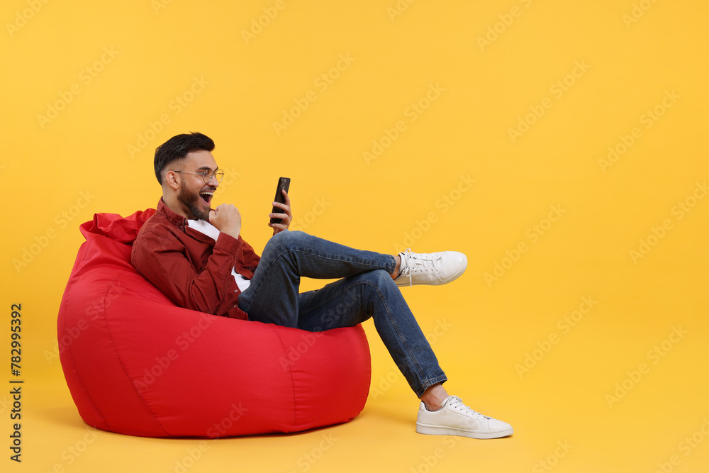 Happy young man using smartphone on bean bag chair against yellow background, space for text Happy young man using smartphone on bean bag chair against yellow background, space for text