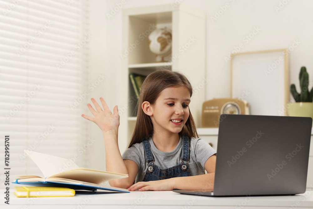 Cute girl waving hello during online lesson via laptop at white table indoors Cute girl waving hello during online lesson via laptop at white table indoors