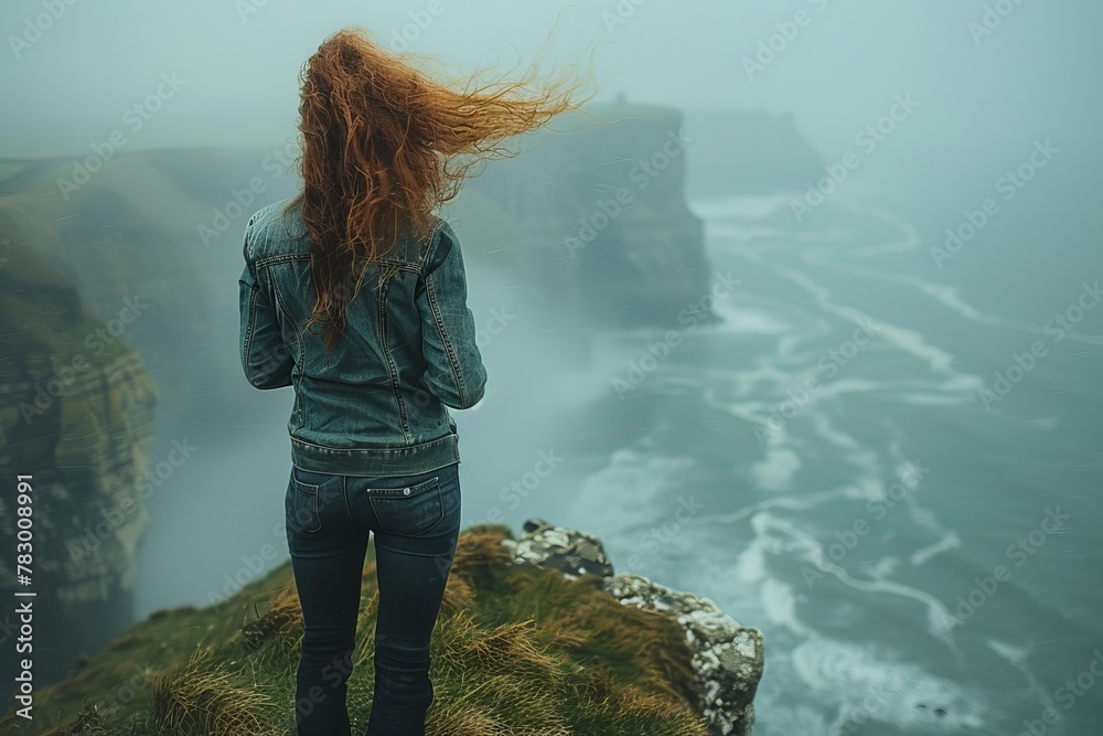 A solitary woman in a denim jacket stands facing the ocean, her hair blowing in the wind atop a mist-covered cliff - obrazy, fototapety, plakaty 