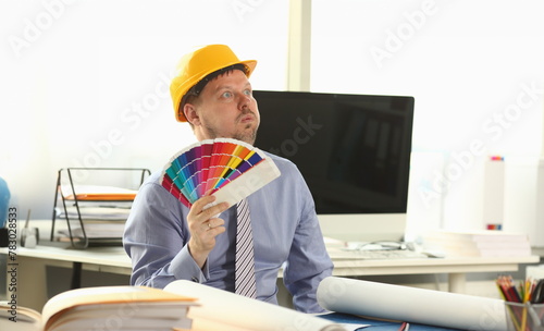 Dreamy Adult Designer Holding Paint Colour Palette. Drafting Tools on Worktop. Building Interior and Creative Technology Concept. Constructor Man in Protective Helmet Sit at Office Workplace