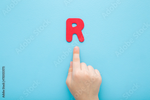 Baby boy hand finger pointing to red R letter on light blue table background. Pastel color. Time to learning. Closeup. Top down view.