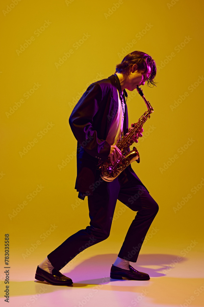 Young virtuoso in classical black suit playing saxophone in neon light against vibrant yellow studio background. Concept of music and art, hobby, concerts and festivals, modern culture. Ad Young virtuoso in classical black suit playing saxophone in neon light against vibrant yellow studio background. Concept of music and art, hobby, concerts and festivals, modern culture. Ad