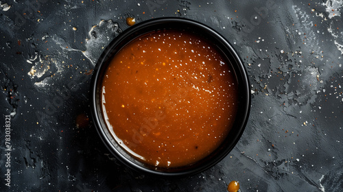 Rich caramel sauce in a black ceramic dish against a textured dark backdrop with vibrant orange splashes. The composition highlights the glossy sheen and smooth consistency photo