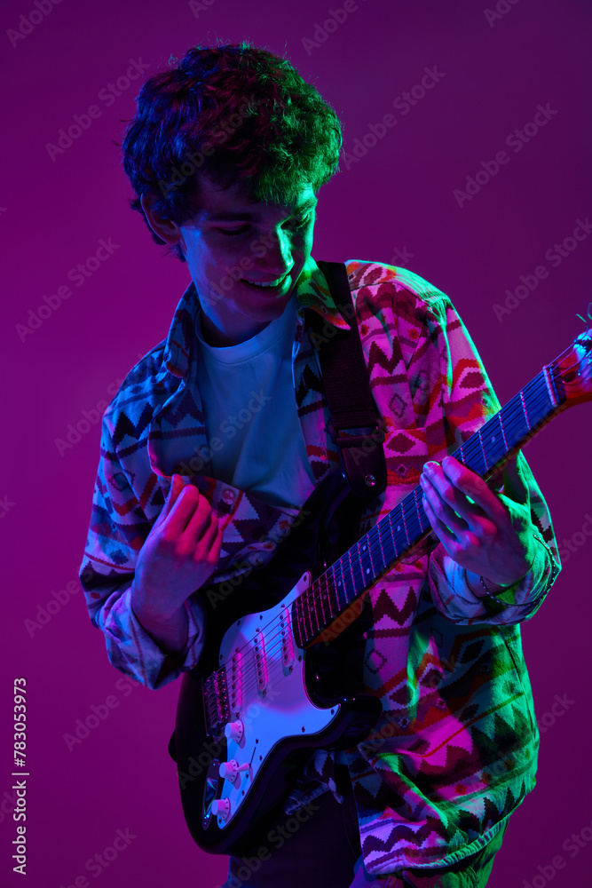 Fototapeta premium Young artistic guitarist in retro outfit performing in pink-purple stage neon light against gradient studio background. Concept of music and art, hobby, concerts and festivals, modern culture. Ad