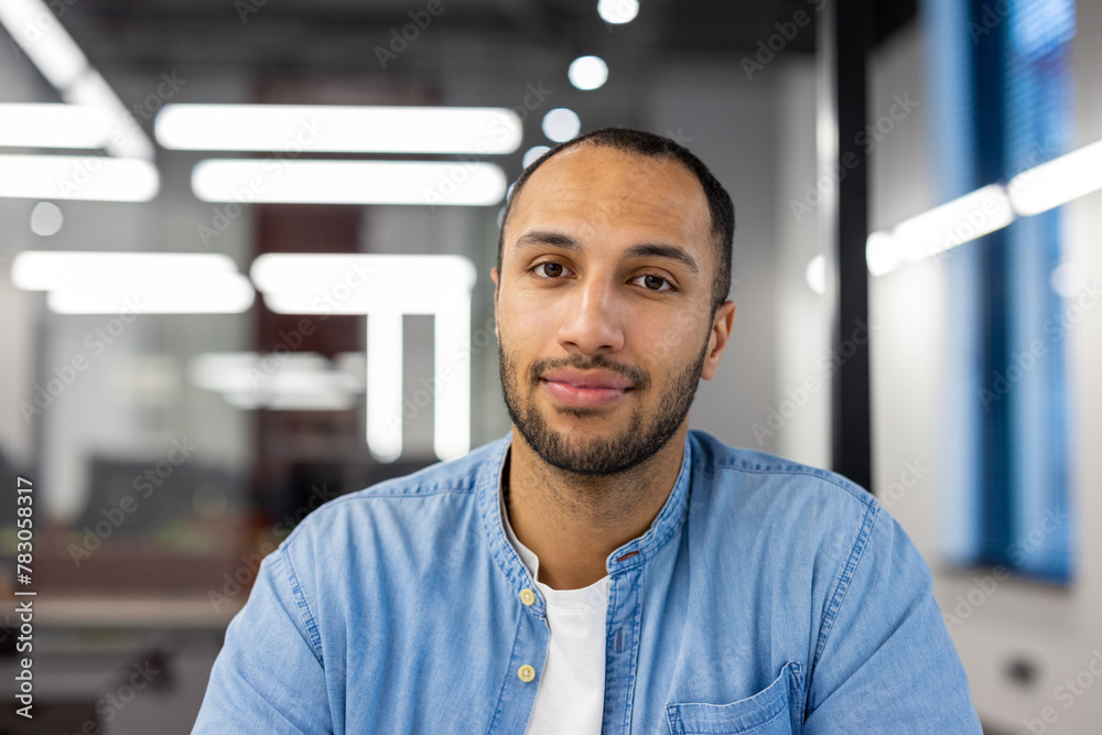 Fototapeta premium Close-up portrait of a young Muslim man sitting in a modern office in a denim shirt and looking confidently at the camera