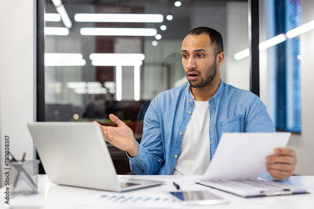Upset young Muslim man sitting in modern office at desk, holding documents in hands and looking at laptop screen in shock, spreading hands Upset young Muslim man sitting in modern office at desk, holding documents in hands and looking at laptop screen in shock, spreading hands