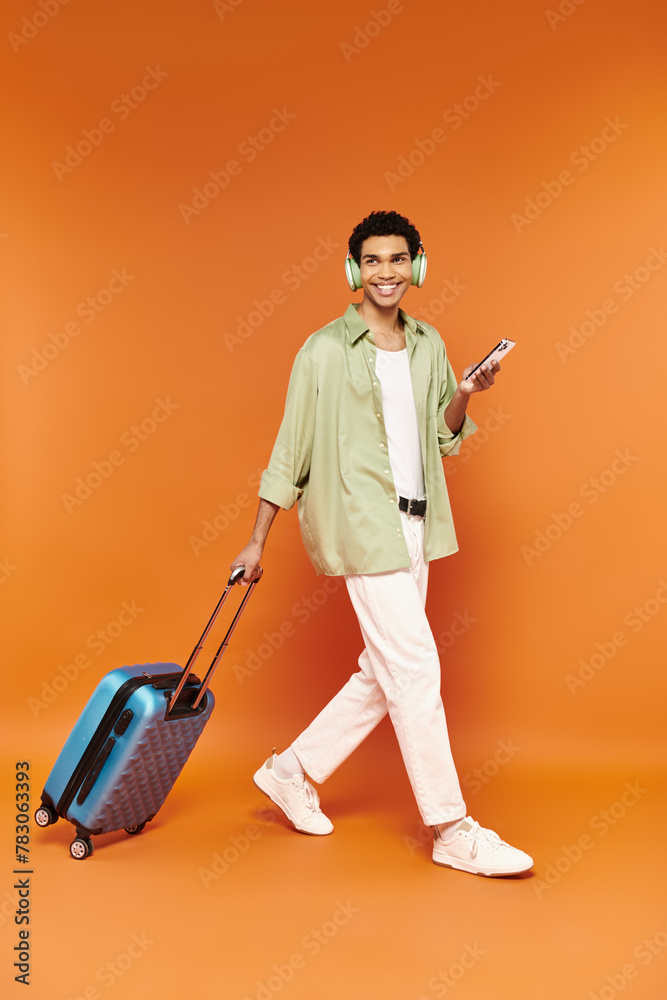 joyous african american man with headphones holding phone and walking with his blue suitcase