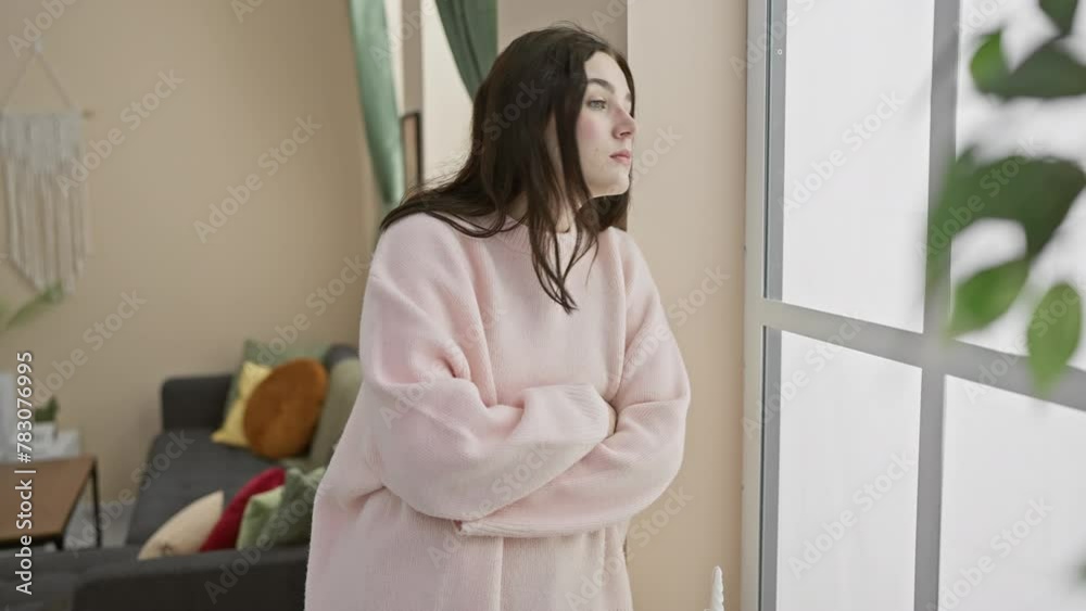 A contemplative young woman in a pink sweater indoors, looking out a window in a cozy, modern living room. A contemplative young woman in a pink sweater indoors, looking out a window in a cozy, modern living room.