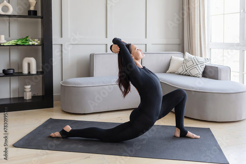 A woman practicing yoga performs a variation of the Ashva Sanchalasana exercise, rider pose, training at home in a black sports overall on a mat, full-length view photo