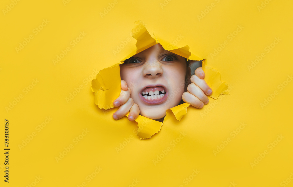 A little girl looks through a torn hole in yellow paper. The concept of anger, fear, fright and shock from what he saw. Unexpected aggression and stress. Copy space. A little girl looks through a torn hole in yellow paper. The concept of anger, fear, fright and shock from what he saw. Unexpected aggression and stress. Copy space.