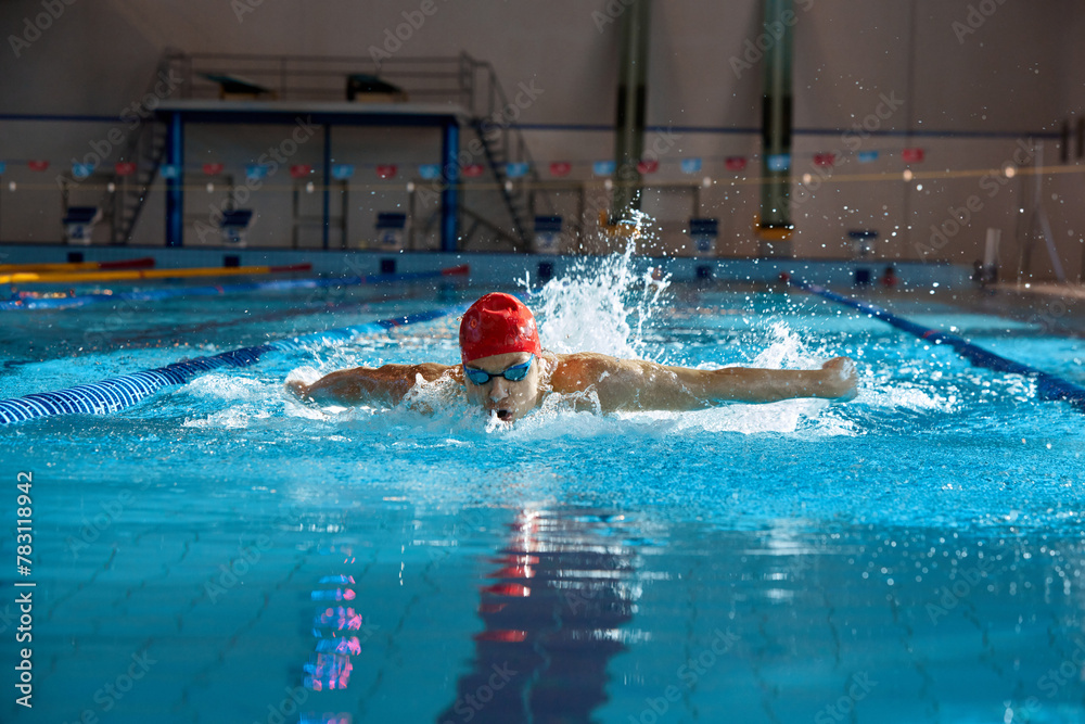 Competitive, muscular young man, swimmer in red cap performs butterfly stroke in pool, training indoors. Concept of professional sport, health, endurance, strength, active lifestyle