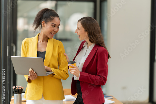 Diverse businesswomen collaborating on a project and talking, using a laptop while sharing ideas and developing strategies in a contemporary office environment