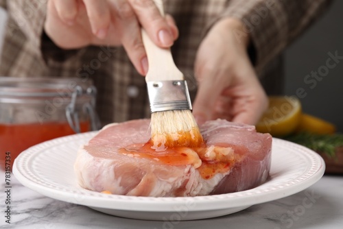 Woman spreading marinade onto raw meat with basting brush at white marble table, closeup