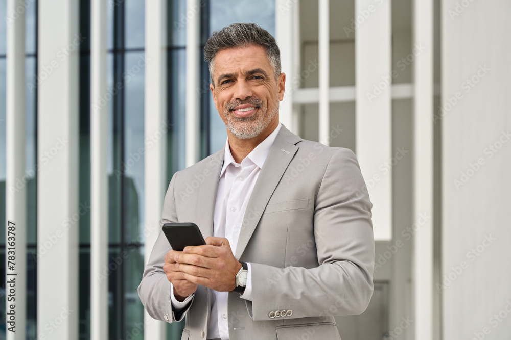 Fototapeta premium Confident happy middle aged Latin banker, smiling mature older business man corporate leader investor entrepreneur wearing suit standing outdoors using smartphone with mobile phone in hands. Portrait