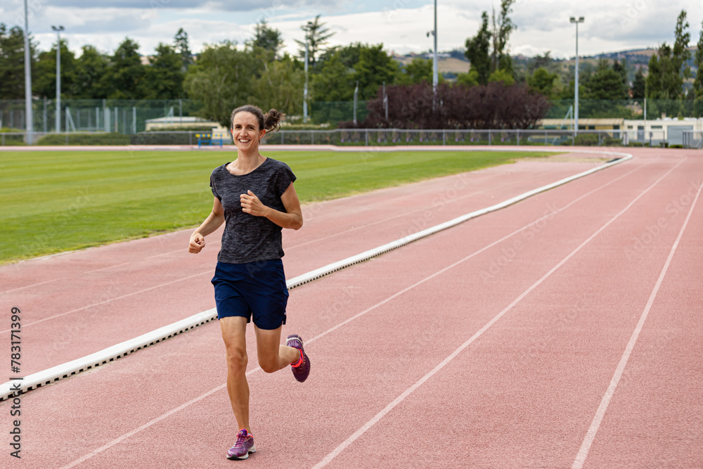 A young woman running on a stadium track, looking at the camera and smiling; laughing female jogger on a track .