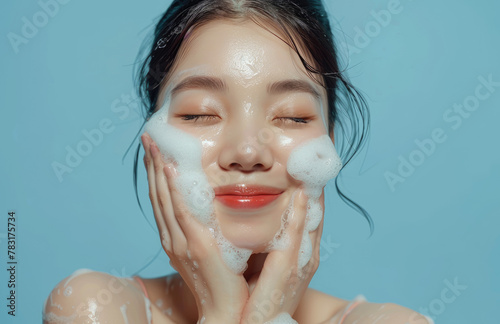 Young korean woman with closed eyes washing her face and smiling while using facial foam on a light blue background