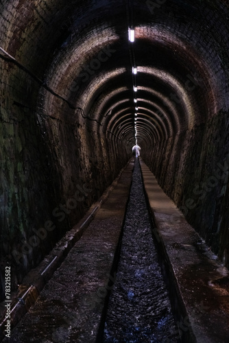 long tunnel, illuminated by artificial lights, surrounded by walls on either side, water channel