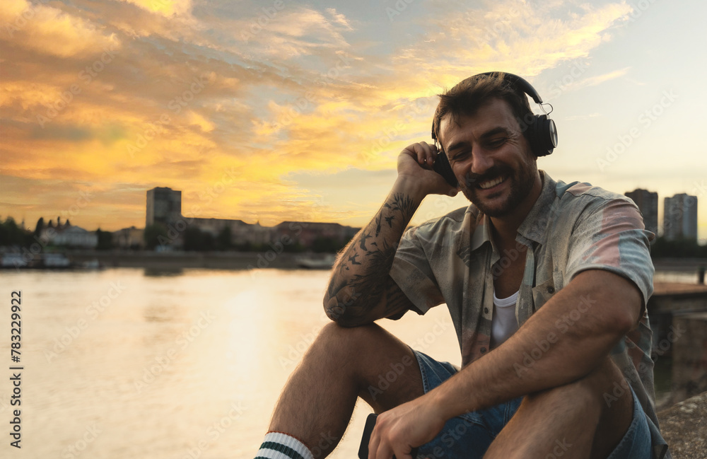 Young hipster millennial man sitting on riverside looking at city listening music on wireless headphones enjoying moment, life and sunset. Happy independent male enjoy freedom and life concept