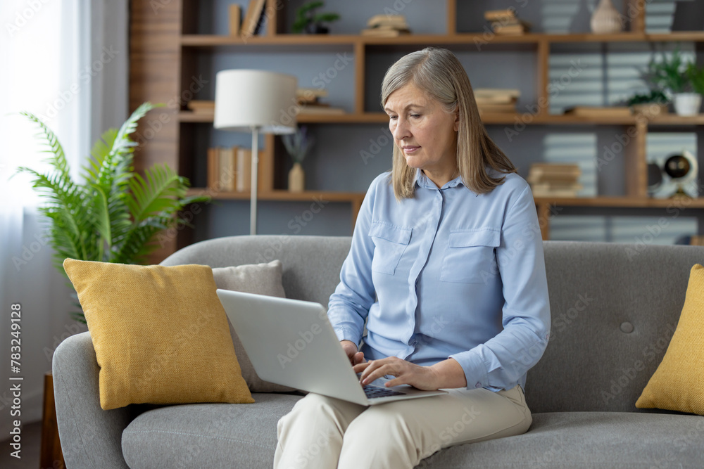 A mature woman, focused and engaged, uses a laptop while sitting on a sofa in a well-decorated living room, suggesting a blend of comfort and productivity. A mature woman, focused and engaged, uses a laptop while sitting on a sofa in a well-decorated living room, suggesting a blend of comfort and productivity.