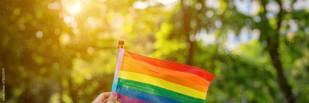 A woman holds the rainbow flag in her hand, outdoors, against green grass and trees, with a blurred background, in a closeup shot of hands holding an LGBT pride symbol, blurred background.
