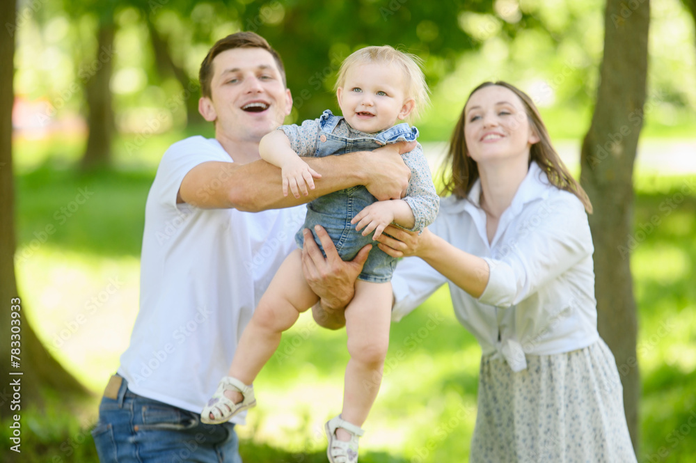 Fototapeta premium Family is having fun outdoors in park: mom and dad swing in arms, throw up child, laugh, feeling happy. Have fun weekend together, pleasant memories, holidays